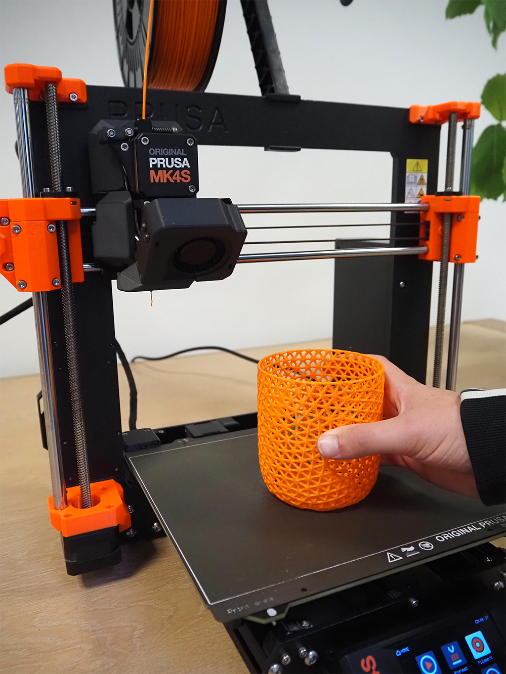PRUSA MK4S 3D printer with an orange cylindrical object printed with BEDROCK 3D PLA on the bed, held by a hand.